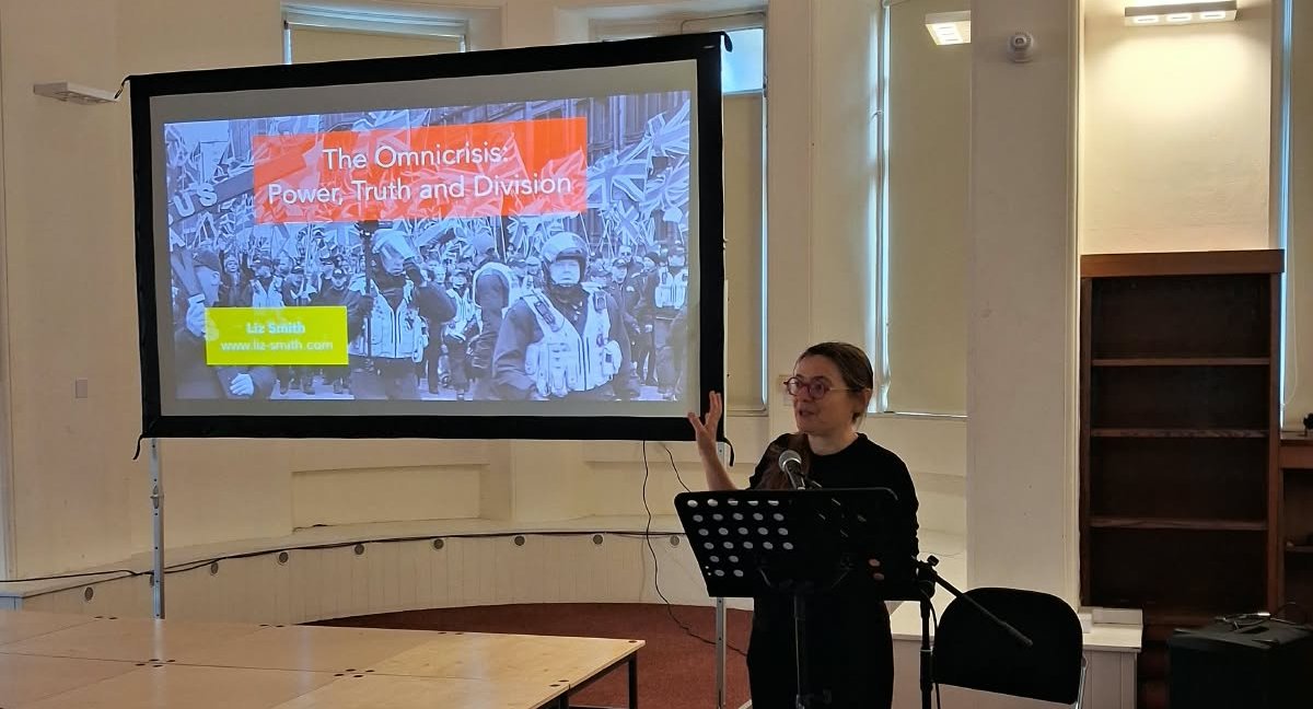 Liz Smith presenting at the lectern beside the title slide at Derby Museum & Art Gallery