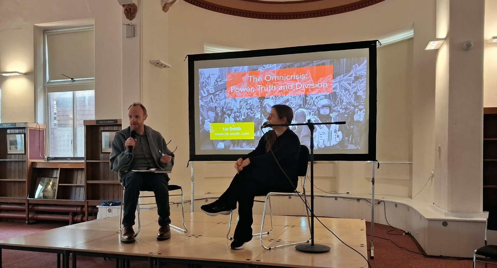 Liz Smith in conversation with Dr Phil Burton-Cartledge on stage at Derby Museum & Art Gallery, beneath a screen showing the title slide